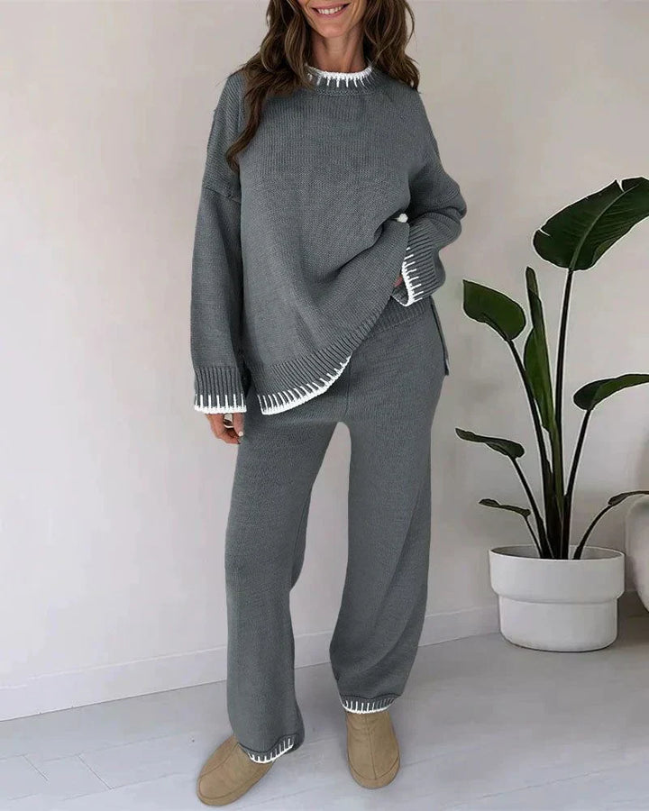 Femme souriante portant un ensemble lounge gris avec finitions blanches côtelées au col, aux poignets et aux ourlets. L’ensemble comprend un pull ample à manches longues et un pantalon droit assorti. Tenue cosy, élégante et confortable, idéale pour un look détente chic à la maison.