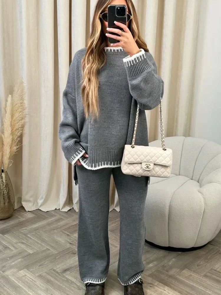 Femme prenant un selfie en miroir, portant un ensemble lounge gris avec finitions blanches contrastantes au col, poignets et ourlets. Le set comprend un pull ample à manches longues et un pantalon droit assorti. Tenue chic, confortable et cosy, accessoirisée d’un sac matelassé beige pour un look raffiné et décontracté.