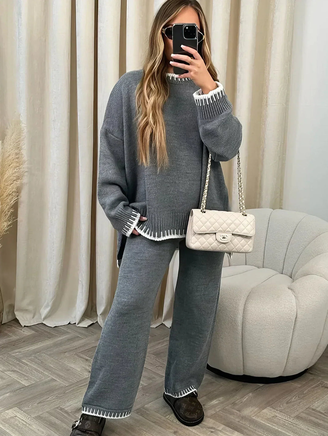 Femme prenant un selfie, vêtue d’un ensemble lounge gris avec finitions côtelées blanches au col, poignets et ourlets. Le set se compose d’un pull oversize à manches longues et d’un pantalon ample assorti. Elle porte un sac matelassé beige et des mocassins, créant un look à la fois chic, confortable et moderne pour l’intérieur ou les sorties décontractées.