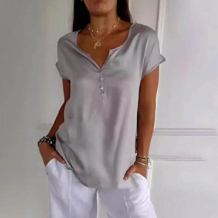 Haut Bliss pour femme, élégant et confortable, coupe fluide à manches courtes, coloris gris avec encolure en V et boutons, porté avec un pantalon blanc pour un style chic et décontracté.