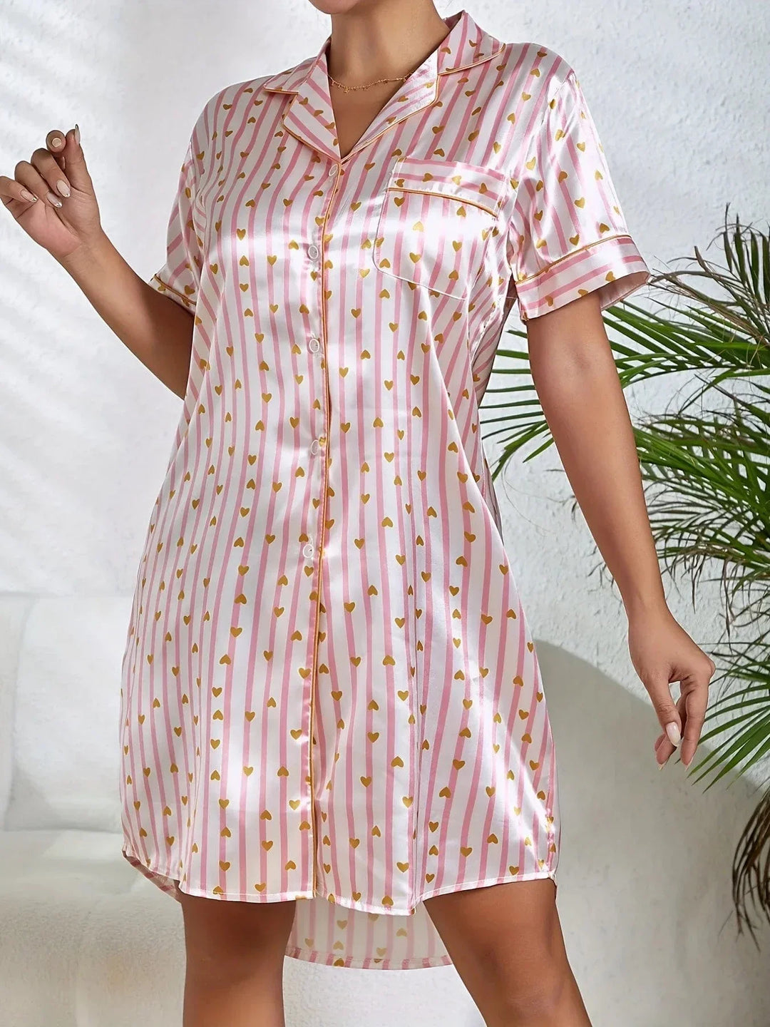Vue avant de la nuisette satinée pour femme, avec un joli motif rayé rose et or orné de cœurs. Elle présente un col classique, une poche sur le côté gauche de la poitrine et des boutons sur toute la longueur. Sa coupe fluide et son tissu satin lui confèrent un style à la fois élégant et confortable, parfait pour une nuit de détente.