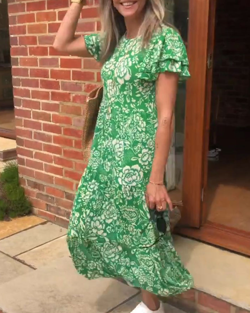 Femme souriante portant une robe verte à fleurs, avec un sac en osier et des baskets blanches, dans une ambiance extérieure ensoleillée.