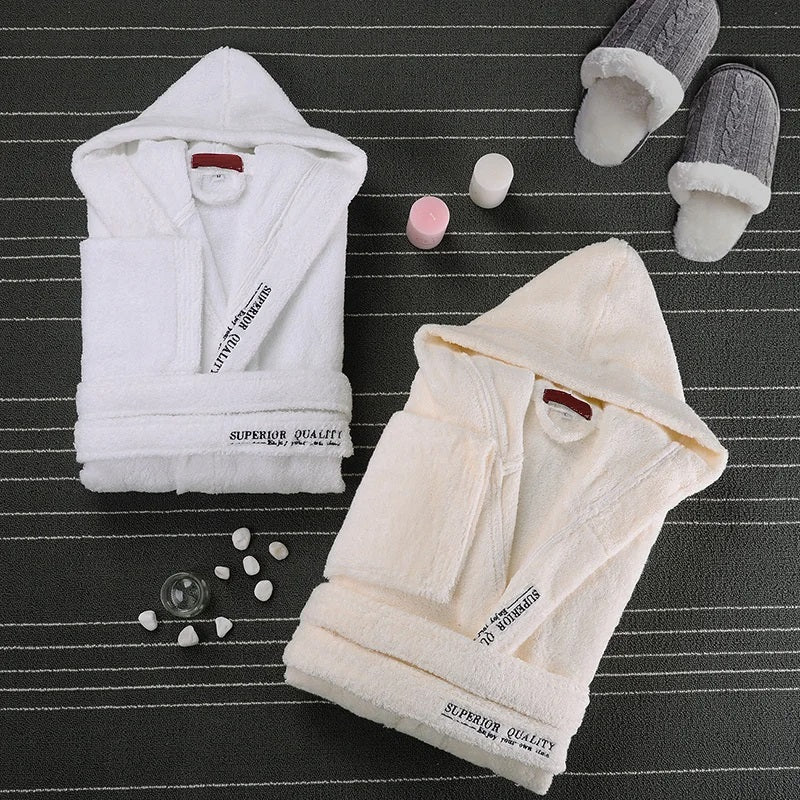 Deux peignoirs Cédric pour homme, modèles doux et relaxants, présentés pliés avec capuche, l’un en blanc et l’autre en beige, accompagnés de chaussons et d’accessoires bien-être sur un tapis sombre.