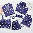 Ensemble de pyjama femme Célia, doux et élégant, composé de plusieurs pièces en satin bleu marine décoré de cœurs blancs : chemise à boutons, débardeur, short, pantalon, bandeau, chouchou et pochette assortie, soigneusement disposés à plat sur un fond clair.