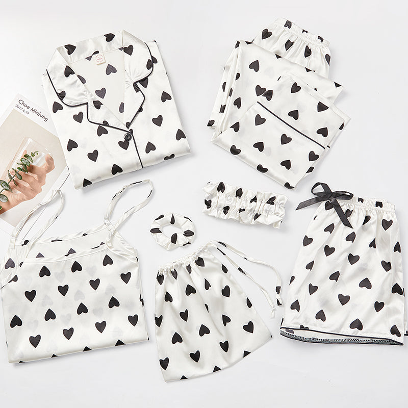 Ensemble de pyjama femme Célia, doux et élégant, composé de plusieurs pièces en satin blanc décoré de cœurs noirs : chemise à boutons, débardeur, short, pantalon, bandeau, chouchou et pochette assortie, soigneusement disposés à plat sur un fond clair.