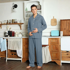 Homme debout dans une cuisine au style rustique, vêtu d’un pyjama bleu ardoise avec passepoils blancs et fermeture boutonnée. Ensemble confortable et raffiné, idéal pour les soirées cocooning ou les matinées détendues à la maison.