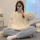 Pyjama femme en peluche, avec un haut blanc à manches longues et un pantalon bleu clair à texture côtelée. La personne prend un selfie tout en portant ce pyjama confortable et stylé, créant une atmosphère décontractée. Le téléphone avec un étui décoré et un sourire joyeux sur une tasse ajoutent une touche de convivialité et de confort à ce moment.