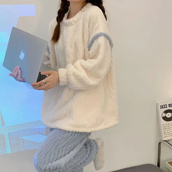 Pyjama femme en peluche avec un haut ample blanc et bleu clair, associé à un pantalon à texture côtelée. La personne tient un ordinateur portable tout en posant dans un décor minimaliste et lumineux. Elle affiche un sourire détendu et porte des vêtements confortables, parfaits pour une journée cosy à la maison.