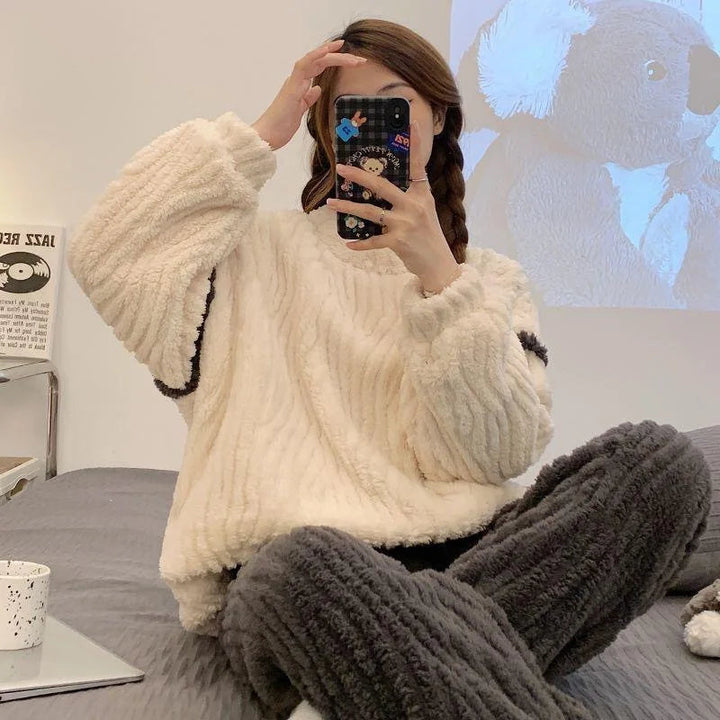 Pyjama tendance pour femme, en tissu pelucheux de couleur crème, avec un haut ample et des pantalons gris foncé à la texture côtelée. La personne, assise sur un lit, prend un selfie en tenant son téléphone et sourit, avec des cheveux en tresses. L&