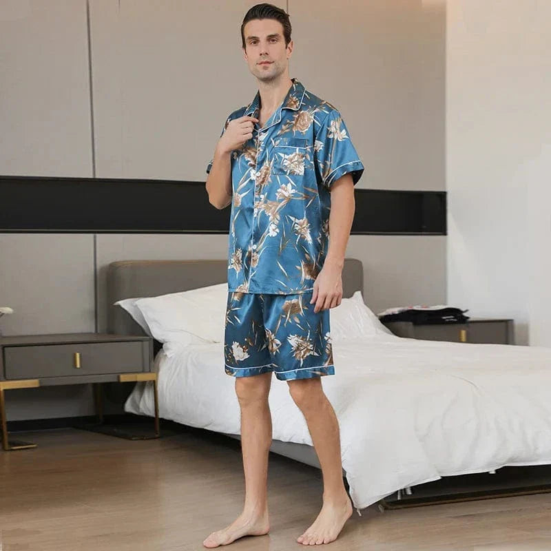 Homme debout portant le pyjama court satin Charles pour homme, ensemble deux pièces avec chemise boutonnée à manches courtes et short assorti, imprimé floral beige sur fond bleu, coupe droite, présenté en intérieur.