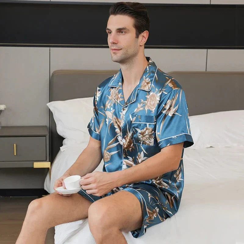 Homme assis portant le pyjama court satin Charles pour homme, ensemble deux pièces avec chemise boutonnée à manches courtes et short assorti, imprimé floral beige sur fond bleu, vue de profil, présenté en intérieur.