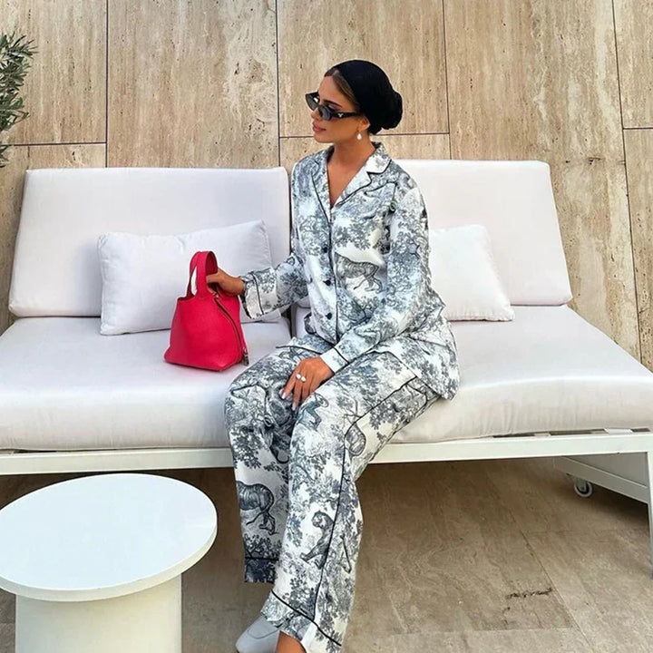 Pyjama femme Charline gris imprimé toile de Jouy, ensemble élégant et confortable deux pièces avec chemise boutonnée à col tailleur et pantalon long assorti, porté assise sur un canapé blanc, coupe fluide et chic.