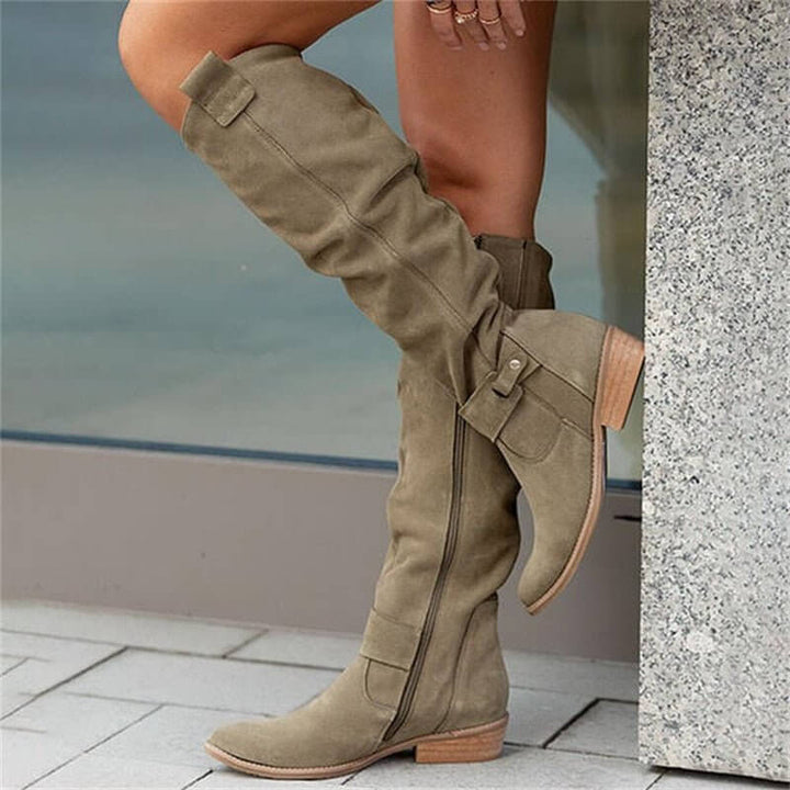 Bottes en daim kaki à tige haute avec une fermeture éclair latérale, parfaites pour un look chic et moderne. Avec des détails de boucles et un talon en bois, elles ajoutent une touche de sophistication et sont idéales pour compléter une tenue stylée et tendance. Confortables et élégantes, ces bottes sont parfaites pour la saison automne-hiver.