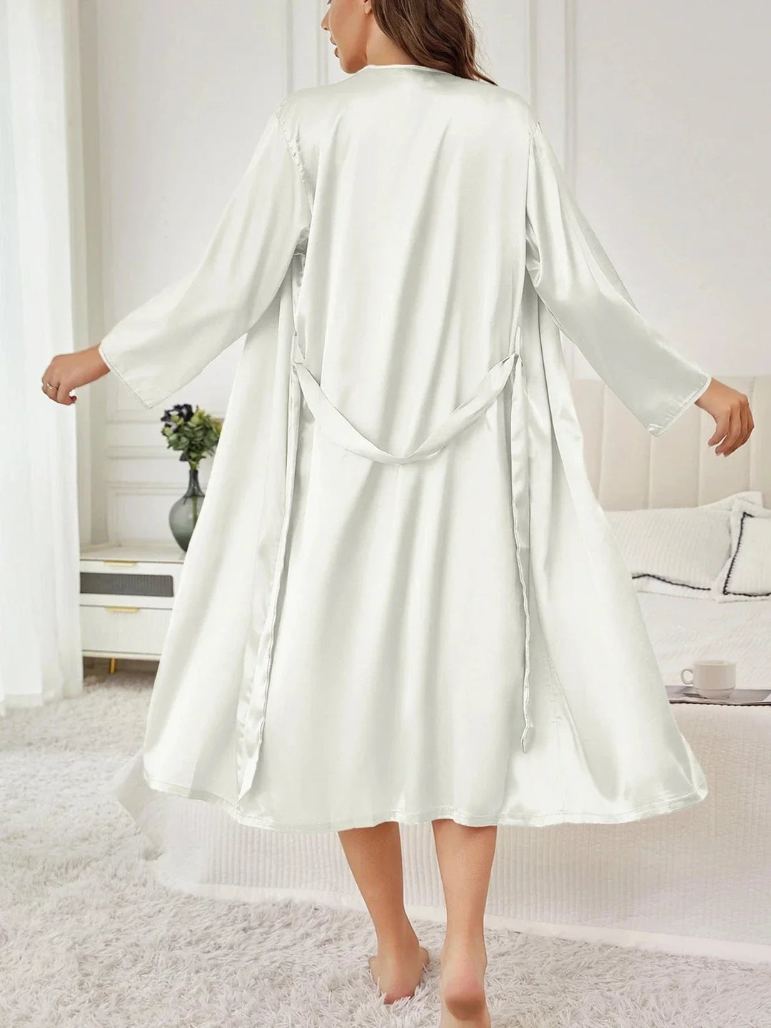 Femme vue de dos marchant pieds nus dans une chambre élégante, portant le peignoir satiné blanc Chloé à manches longues et coupe fluide, avec ceinture à nouer, illustrant la légèreté et le confort raffiné de ce vêtement de nuit féminin.
