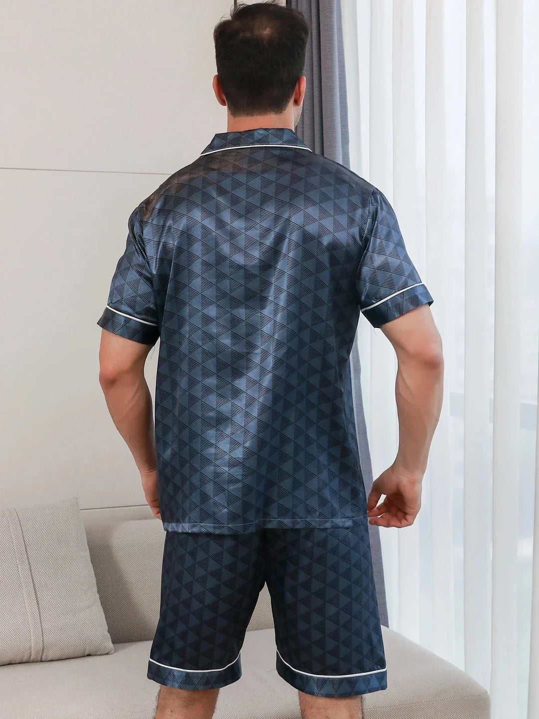 Vue arrière d’un homme portant le shorty satin Christophe pour homme, ensemble deux pièces avec chemise à manches courtes et short assorti, motif géométrique bleu foncé, coupe droite, présenté en intérieur.