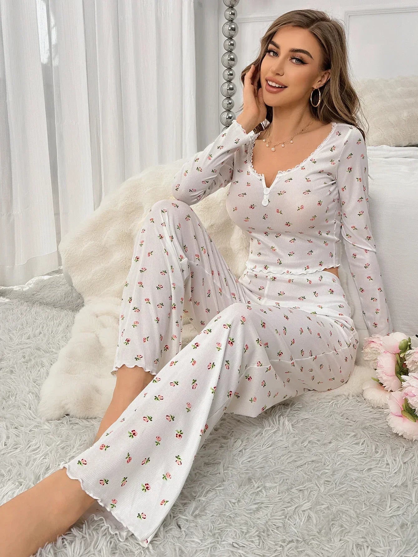 Pyjama floral femme Clarisse blanc, modèle assis au bord du lit portant un ensemble deux pièces doux et confortable avec haut à manches longues, col en V boutonné et bordures festonnées, pantalon large imprimé petites fleurs, coupe décontractée.