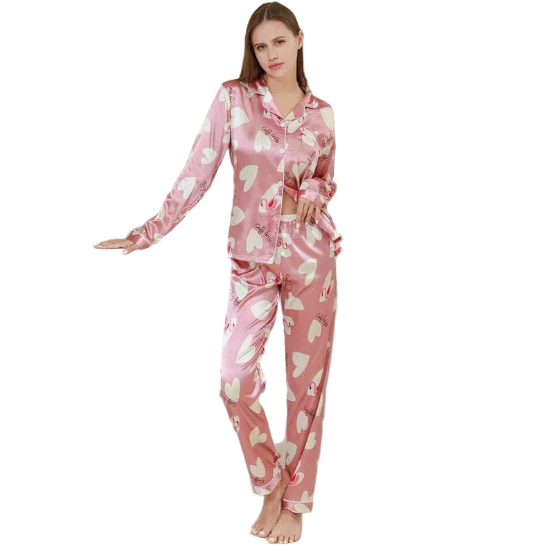 Femme portant un pyjama en satin rose orné de motifs cœurs blancs et d'inscriptions romantiques. Ensemble deux pièces avec chemise à manches longues boutonnée et pantalon assorti. Pyjama tendance, doux au toucher et agréable à porter, parfait pour des nuits stylées et confortables.