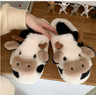 Chaussons femme Cyndelle doux et antidérapants en forme de vache, avec cornes et oreilles en peluche, posés sur une table en bois et tenus par une main.