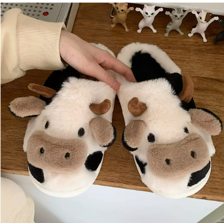 Chaussons femme Cyndelle doux et antidérapants en forme de vache, avec cornes et oreilles en peluche, posés sur une table en bois et tenus par une main.