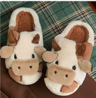 Chaussons femme Cyndelle doux et antidérapants en peluche, motif vache marron et blanc avec cornes et oreilles, posés sur un plaid à carreaux.