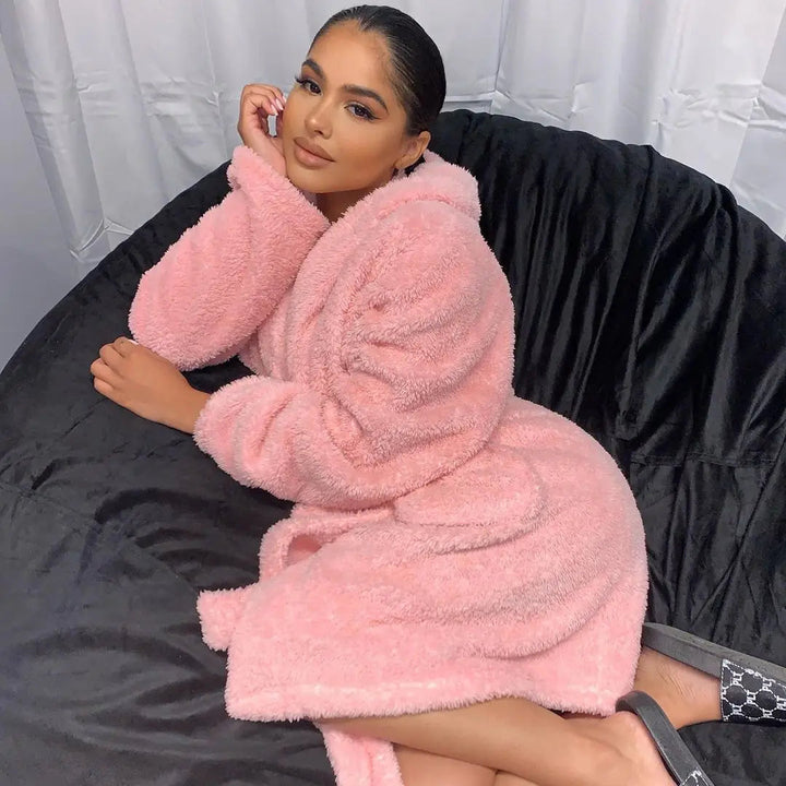 Femme portant le peignoir en coloris rose, un modèle long à la texture ultra-douce et chaude, parfaite pour l’hiver. Elle est confortablement installée sur un fauteuil, illustrant le côté cocooning du peignoir.