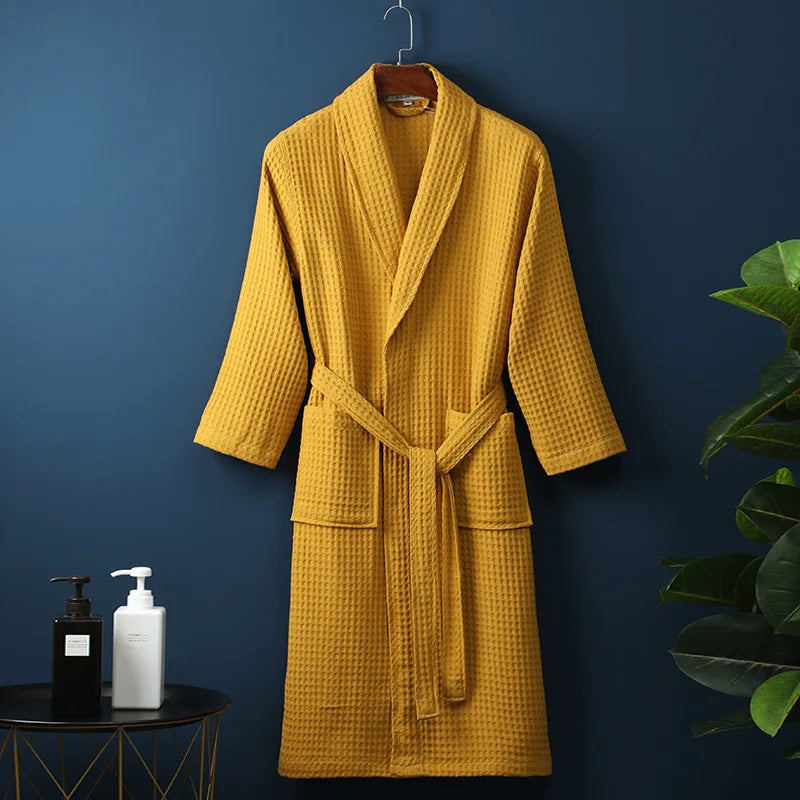 Peignoir Damien pour homme, modèle moelleux et confortable en tissu gaufré jaune moutarde, présenté suspendu devant un mur bleu avec accessoires de salle de bain en décor.