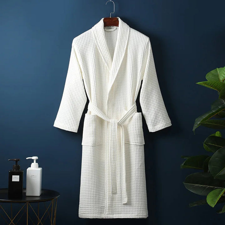 Peignoir Damien pour homme, modèle moelleux et confortable en tissu gaufré blanc, présenté suspendu devant un mur bleu avec accessoires de salle de bain en décor.