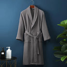 Peignoir Damien pour homme, modèle moelleux et confortable en tissu gaufré gris, présenté suspendu devant un mur bleu avec accessoires de salle de bain en décor.