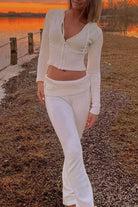 Femme marchant au bord d’un lac au coucher du soleil, vêtue de l’ensemble Dani en maille blanche, composé d’un haut zippé à manches longues et d’un pantalon taille basse assorti. Tenue douce, féminine et élégante, idéale pour un look raffiné et confortable en extérieur.