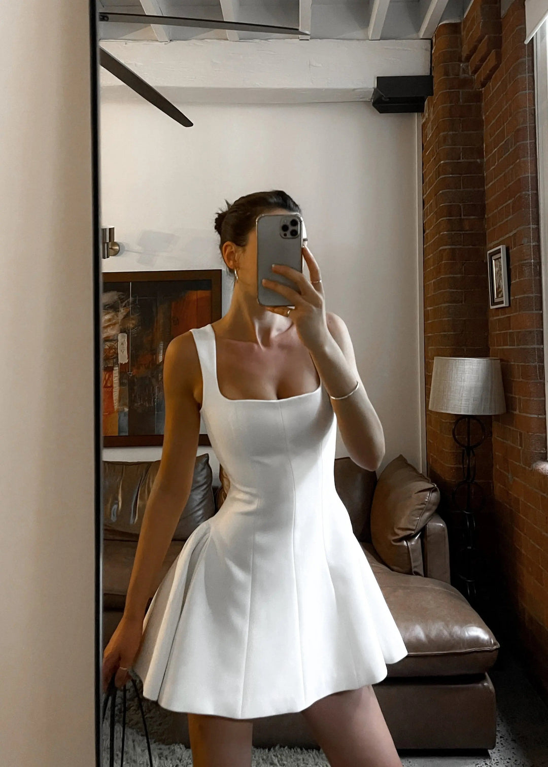 Robe mini blanche Delilah à encolure carrée, coupe cintrée avec jupe évasée, portée lors d’un selfie miroir dans un intérieur moderne une tenue élégante et tendance qui sublime la silhouette.