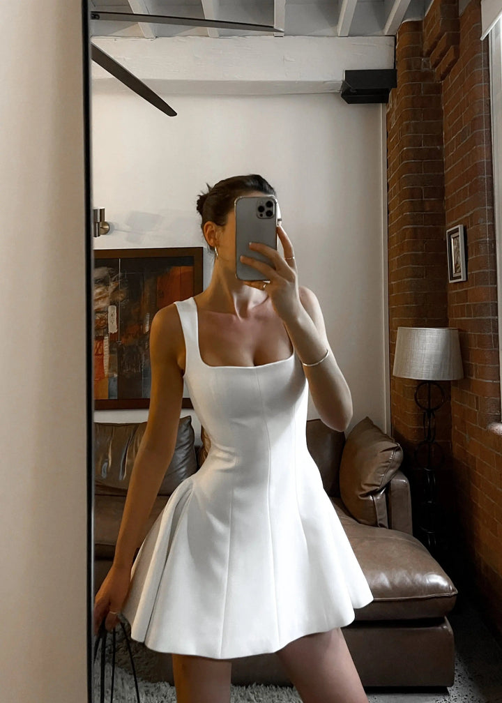 Robe mini blanche Delilah à encolure carrée, coupe cintrée avec jupe évasée, portée lors d’un selfie miroir dans un intérieur moderne une tenue élégante et tendance qui sublime la silhouette.