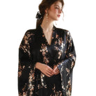 Femme portant le peignoir Denise en satin noir avec motifs floraux dorés et rosés, coupe fluide à manches longues et col en V. Modèle élégant et agréable, parfait pour une allure sophistiquée et féminine à la maison.