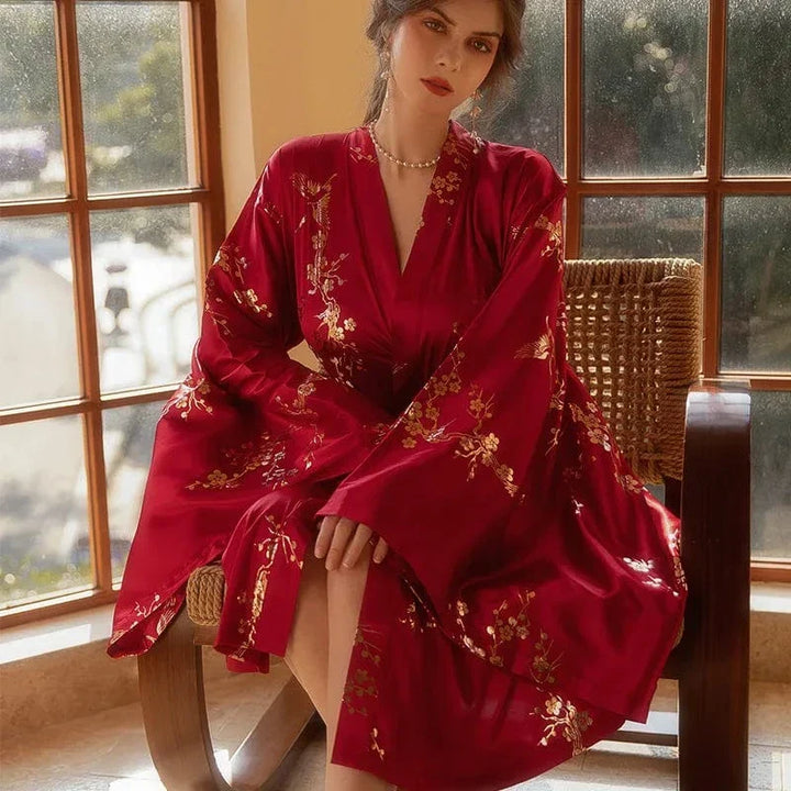 Femme assise près d’une fenêtre, portant le peignoir Denise en satin rouge orné de motifs floraux dorés. Coupe fluide avec manches longues évasées et col en V. Modèle raffiné et confortable, idéal pour une allure élégante à la maison.