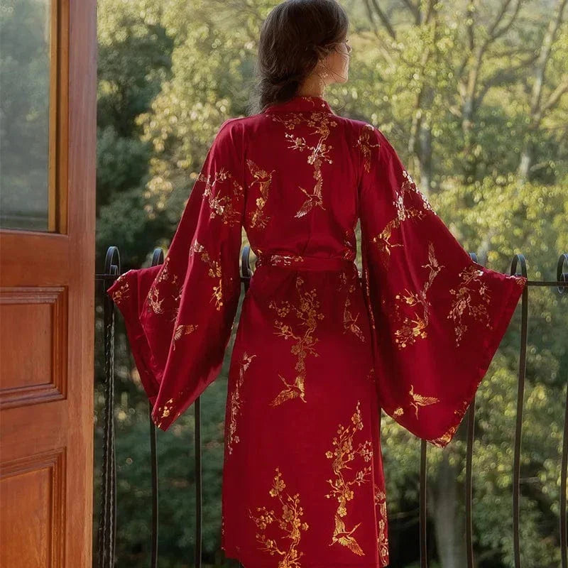 Vue de dos d’une femme portant le peignoir Denise en satin rouge avec motifs dorés floraux et oiseaux, manches longues évasées et ceinture nouée à la taille. Modèle élégant et fluide, parfait pour une touche de raffinement au quotidien.