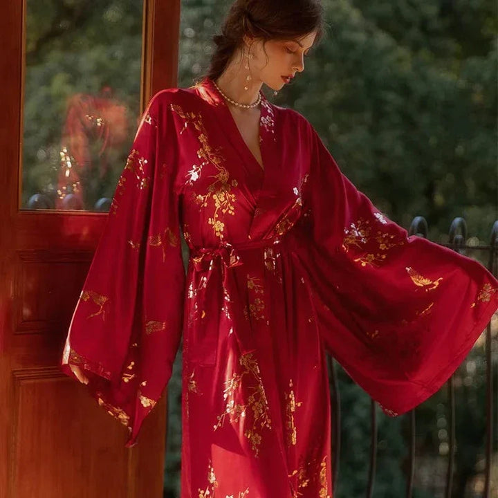 Femme debout près d’une porte ouverte, portant le peignoir Denise en satin rouge orné de motifs dorés floraux et d’oiseaux. Manches longues amples et ceinture nouée à la taille. Modèle raffiné et agréable, parfait pour une élégance douce à la maison.