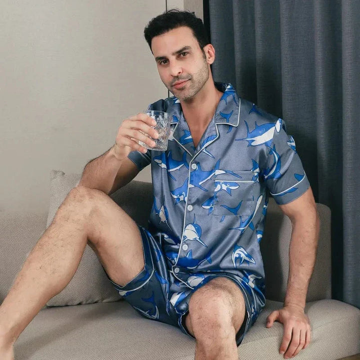 Homme assis sur un canapé portant le shorty satin pour homme Edouard, ensemble tendance et confortable composé d’une chemise à manches courtes et d’un short assorti, en satin gris-bleu à imprimé requins, offrant un style moderne, décontracté et un confort idéal pour la détente à la maison.