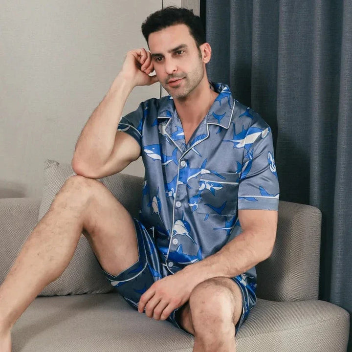 Homme assis sur un canapé portant le shorty satin pour homme Edouard, ensemble tendance et confortable en satin gris-bleu à imprimé requins, composé d’une chemise à manches courtes boutonnée et d’un short assorti, offrant une allure moderne et un confort idéal pour la détente à la maison.
