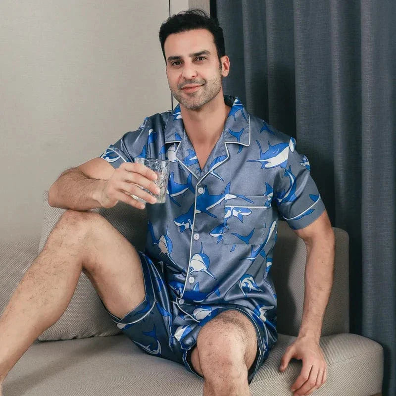 Homme assis sur un canapé tenant un verre, portant le shorty satin pour homme Edouard, ensemble tendance et confortable composé d’une chemise à manches courtes et d’un short assorti, en satin gris-bleu à imprimé requins, offrant un style moderne et un confort idéal pour la détente à la maison.