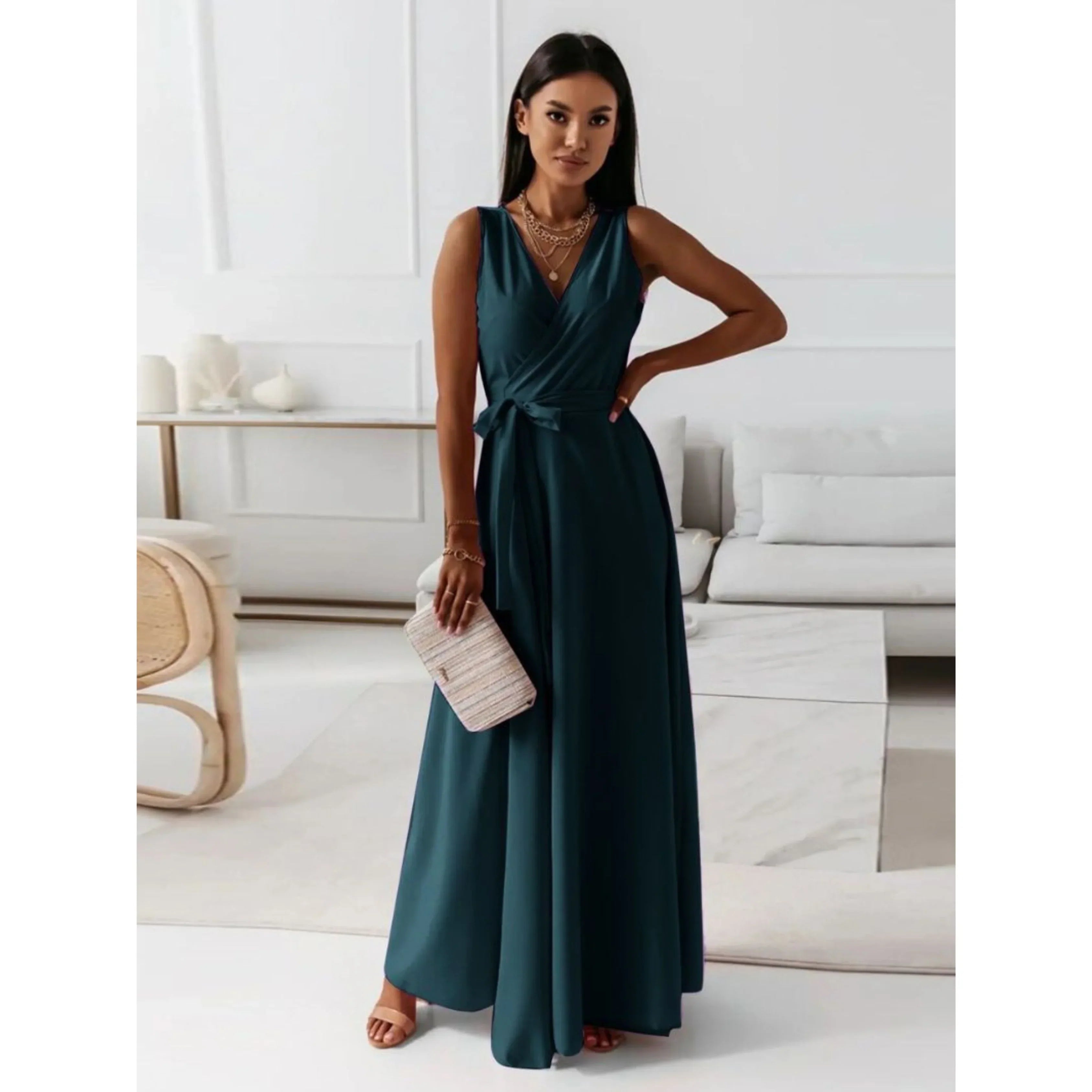 Vert émeraude foncé, décolleté en V croisé, taille ceinturée et jupe fluide, avec sandales et pochette, dans un intérieur lumineux.