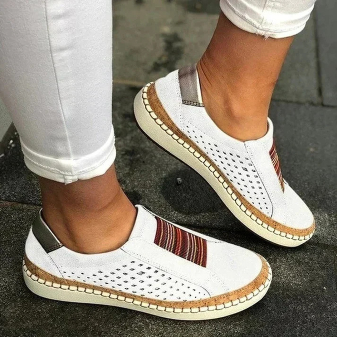 Chaussures femme confortables et idéales au quotidien, slip-on blanches avec bande rayée, perforations respirantes et semelle épaisse beige, portées avec un pantalon blanc.