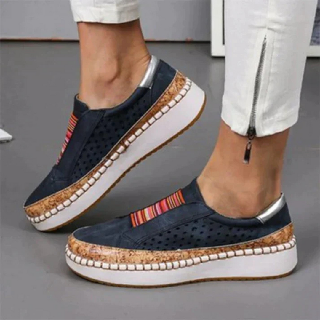 Chaussures femme confortables et idéales au quotidien, slip-on bleu marine avec bande rayée, perforations respirantes et semelle compensée blanche avec liseré effet liège, portées avec un pantalon blanc zippé.