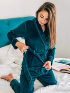 Femme à genoux sur un lit portant un pyjama en velours vert foncé composé d’une veste croisée à ceinture et d’un pantalon assorti. Ensemble chaud, doux et très confortable, idéal pour l’hiver et les moments cocooning à la maison.
