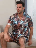 Homme assis portant le pyjama satin Enzo pour homme, ensemble deux pièces avec chemise à manches courtes boutonnée et short assorti, imprimé feuillage multicolore, vu de profil, présenté en intérieur.