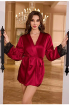 Kimono femme Évangéline en satin bordeaux, coupe courte avec ceinture à nouer, manches longues finies de dentelle noire, porté debout dans un intérieur élégant avec lustre en arrière-plan.