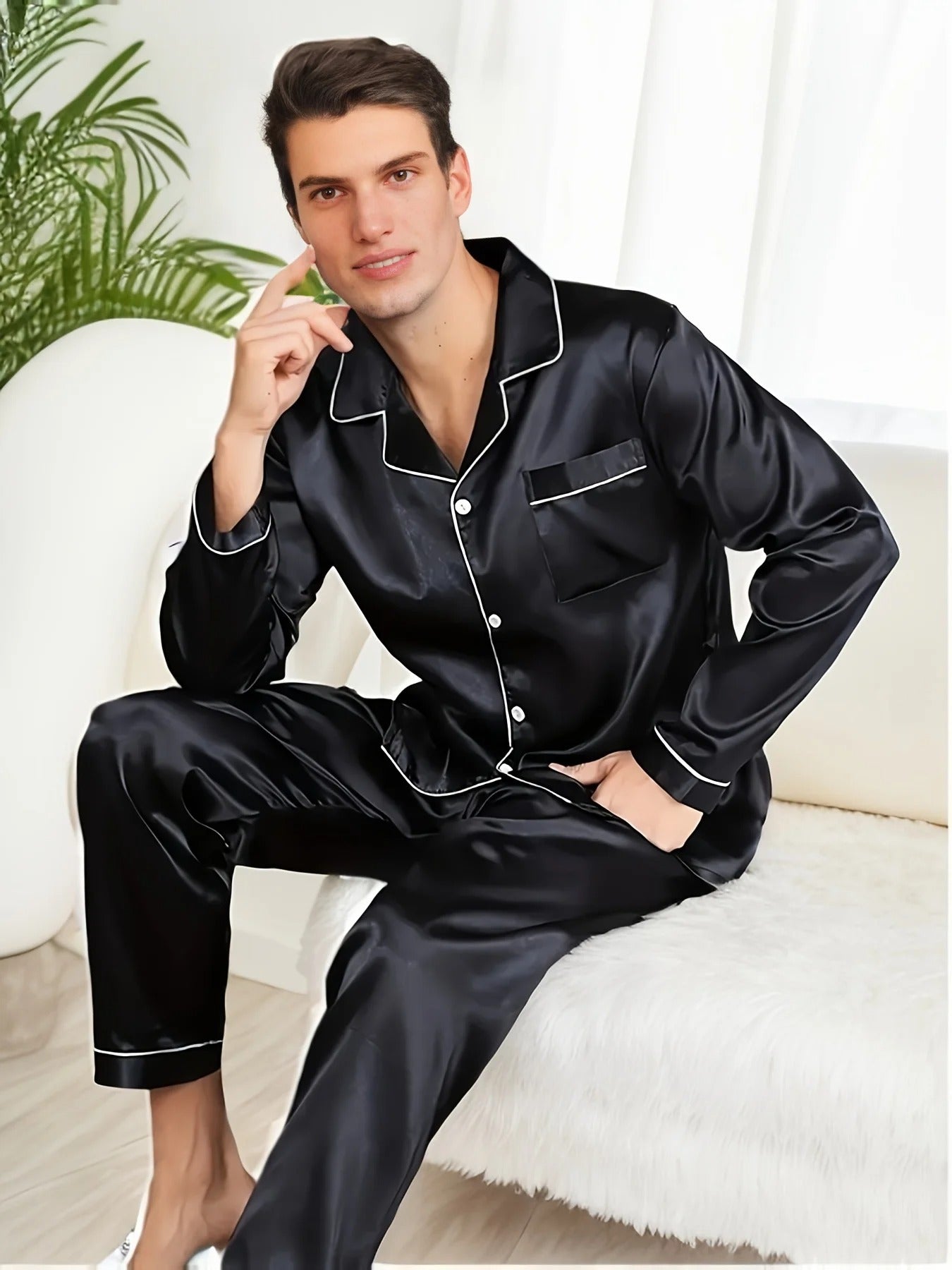 Homme assis sur un canapé, portant un pyjama noir en satin avec chemise boutonnée à col et passepoils blancs, accompagné d’un pantalon assorti. Ensemble élégant, doux et confortable, idéal pour les soirées relax et le sommeil tout en style.