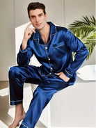Homme assis en tenue d’intérieur, portant un pyjama long en satin bleu nuit, composé d’une chemise boutonnée à col et passepoils blancs, avec une poche poitrine, et d’un pantalon assorti. Ensemble élégant, doux et confortable, parfait pour les soirées cocooning ou les nuits raffinées.