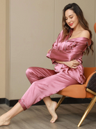 Femme assise dans un fauteuil, souriante et détendue, portant le pyjama Felicity en satin rose framboise avec passepoils clairs. Ensemble deux pièces raffiné et soyeux, alliant élégance, confort et féminité pour les soirées cocooning.
