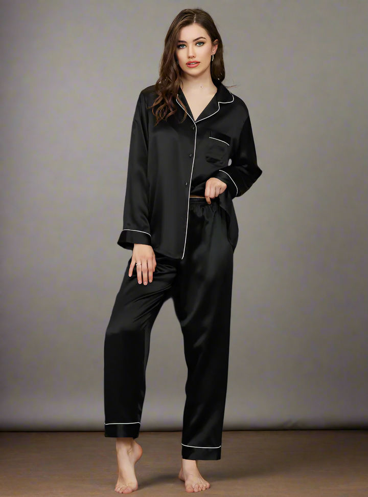 Femme debout portant le pyjama Felicity en satin noir avec passepoils blancs. Ensemble deux pièces chic et fluide, à la fois doux et élégant, idéal pour des nuits raffinées ou des moments de détente avec style.
