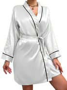 Femme portant un peignoir court en satin blanc à finitions noires, avec ceinture à nouer à la taille et col châle. Peignoir doux, élégant et léger, idéal pour les instants de détente ou pour une touche chic au quotidien.