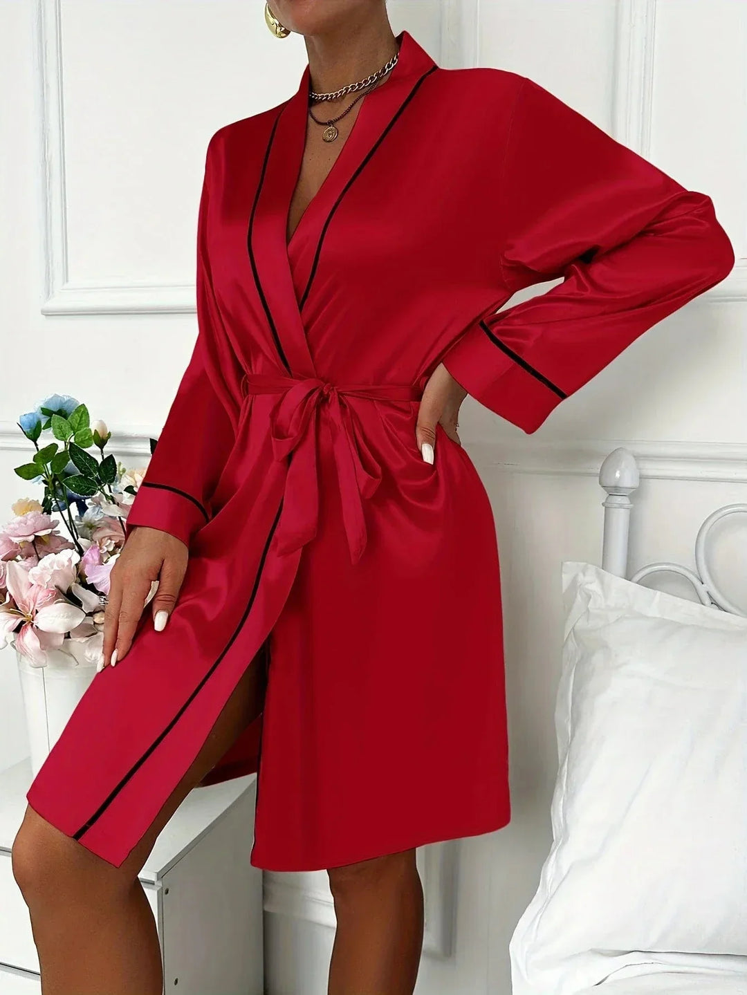 Femme portant un peignoir court en satin rouge vif, agrémenté de liserés noirs élégants, avec ceinture à nouer et col châle. Modèle raffiné et soyeux, parfait pour des moments de détente glamour ou une allure romantique à la maison.